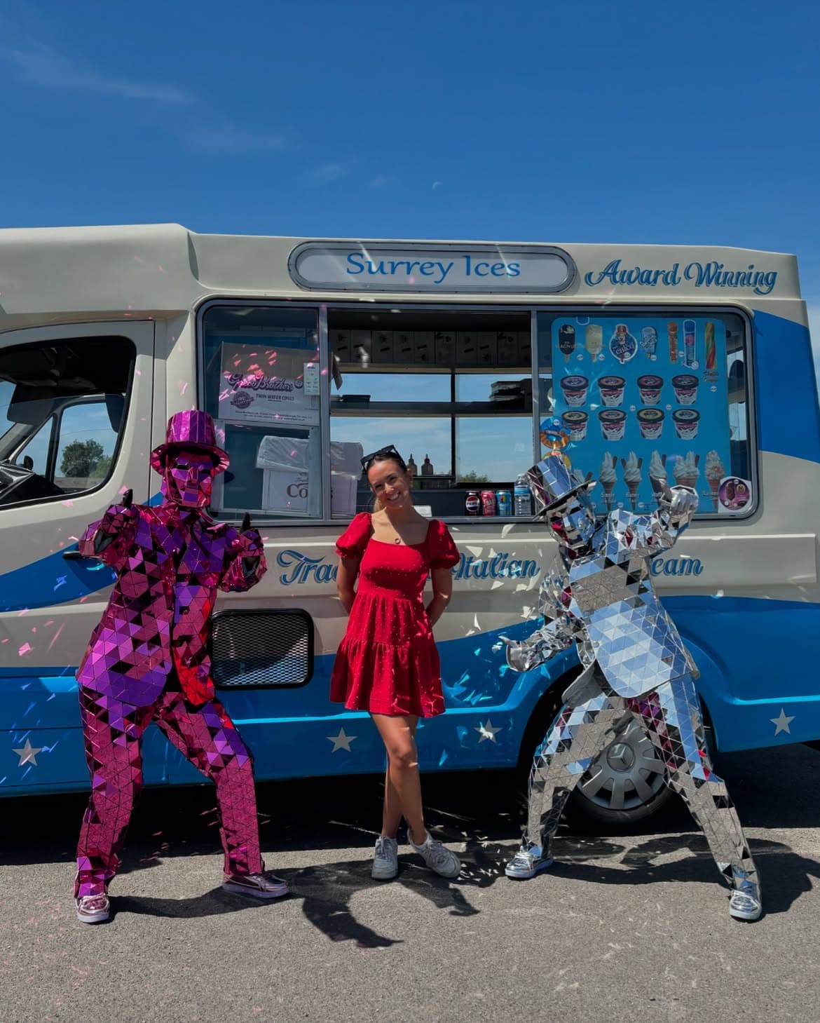 A woman in a red dress standing between two mirrored costumes in front of a blue and cream Surrey Ices van