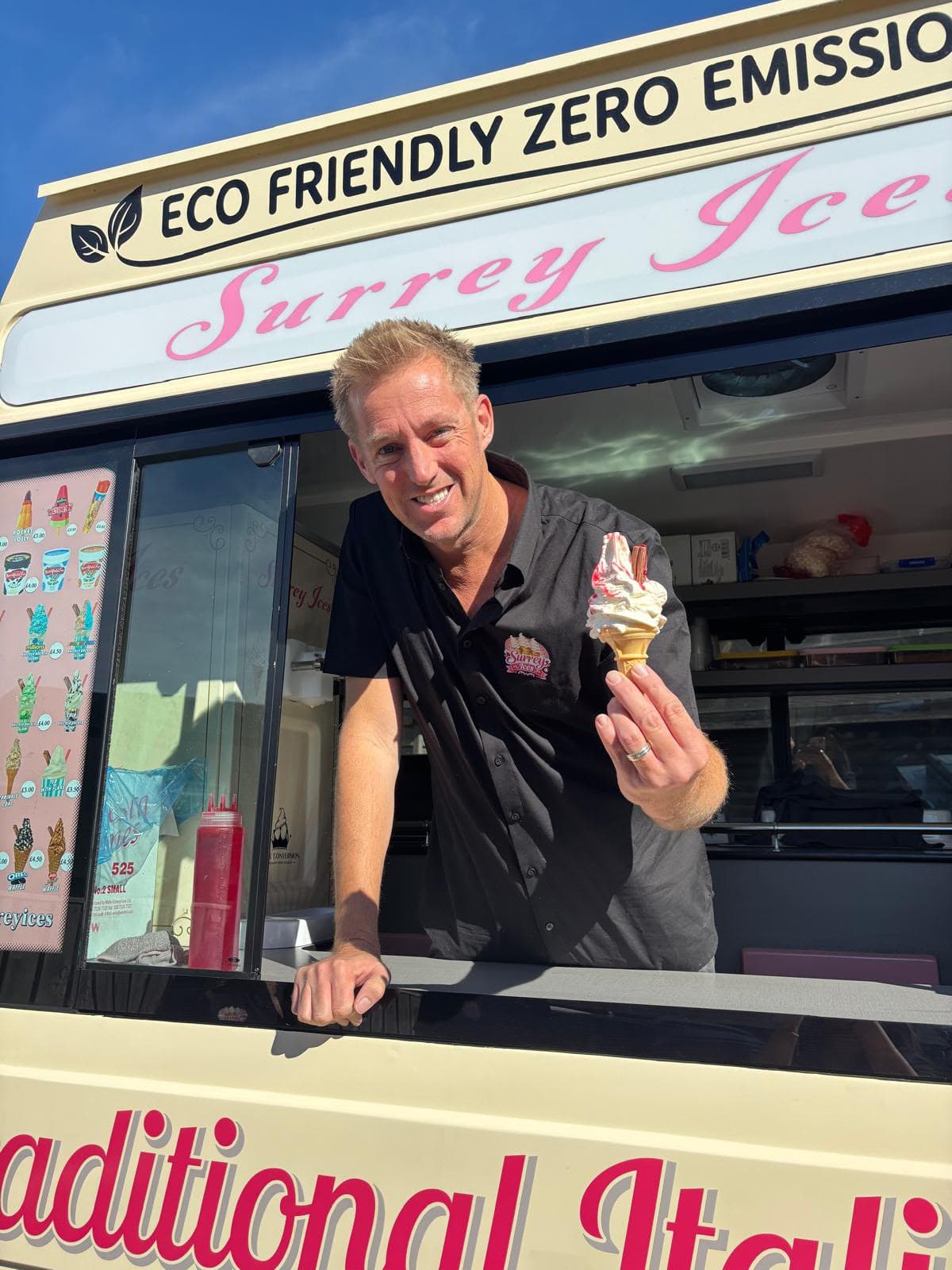 A Surrey Ices server leaning from the hatch of an eco-friendly van, holding a soft serve cone