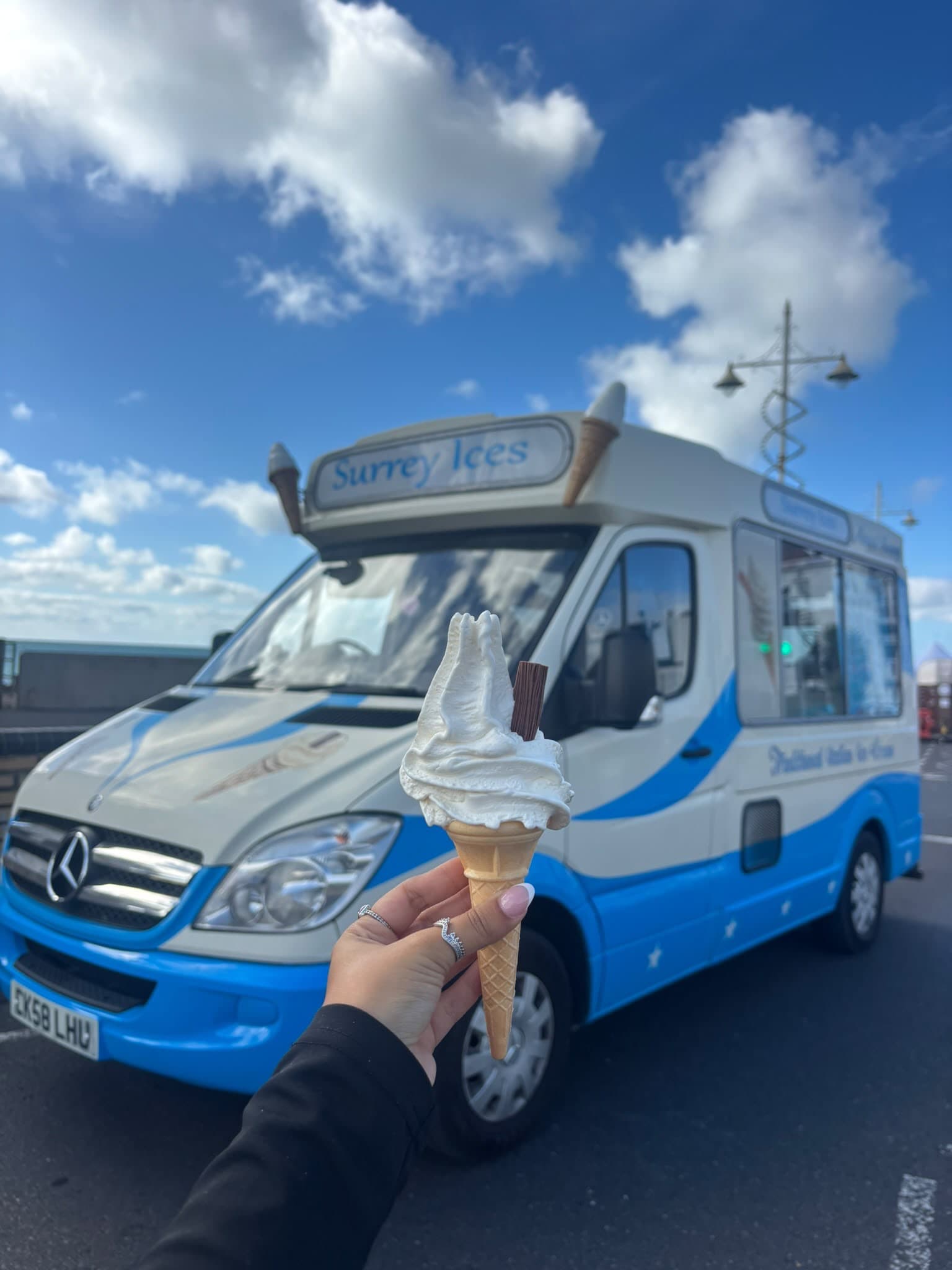A hand holding a soft serve cone with a chocolate flake in front of a blue and cream Surrey Ices van