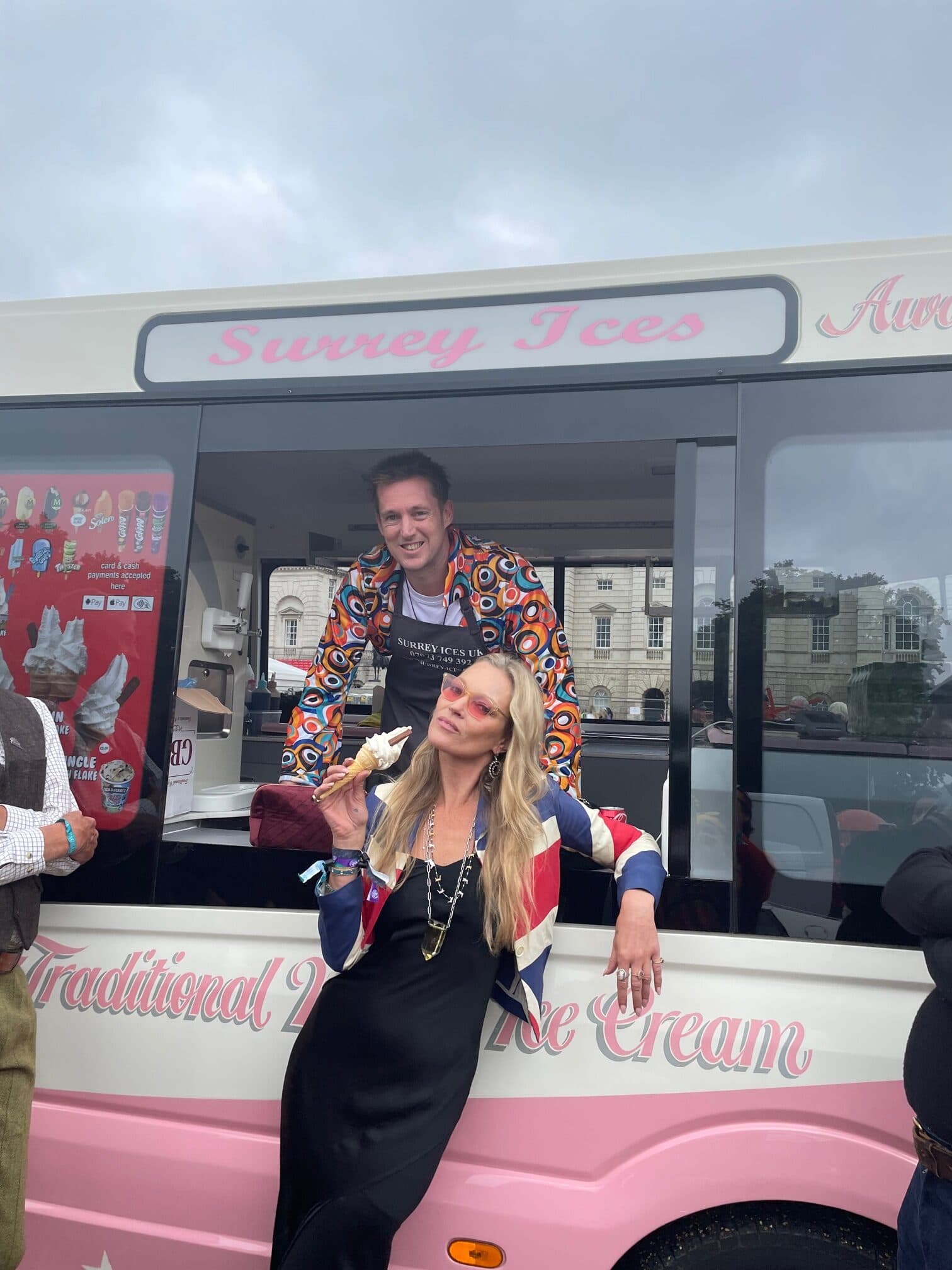 A woman in a Union Jack jacket holding a cone beside a cream and pink Surrey Ices van