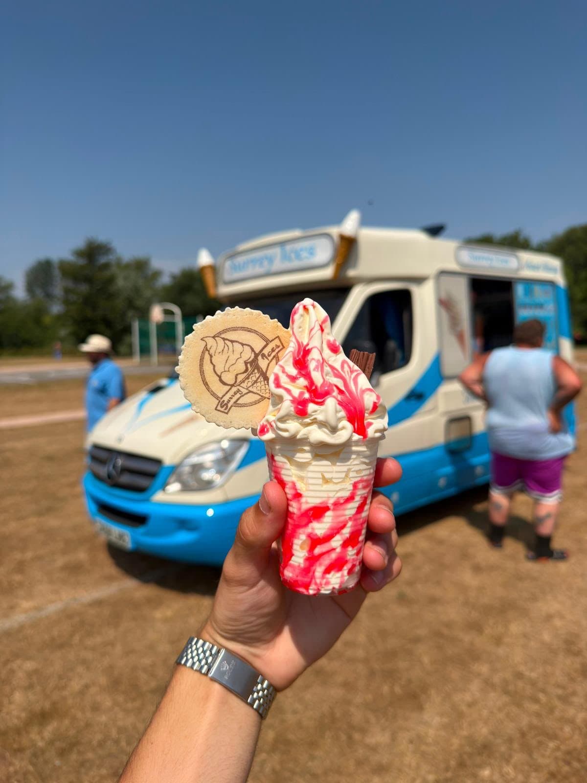 A hand holding a soft serve cone with red sauce and a Surrey Ices wafer, with a blue and cream van on a playing field behind